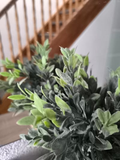 The image shows a close-up view of a green plant, likely an artificial one given the uniform texture and slight dustiness on its leaves. The plant is predominantly in the foreground and mid-ground, with its leaves ranging in color from a lighter, almost neon green to a darker, muted green. Some leaves appear lighter and newer, while others are darker and more established, creating a layered effect.
In the background, a wooden staircase with vertical balusters is visible, suggesting an indoor setting, possibly a home or a commercial space. The staircase has a warm, natural wood tone. To the right of the staircase, a light-colored wall is present. The overall lighting suggests it could be daytime, as there is ambient light illuminating the scene, but no direct sunlight is apparent. The focus is primarily on the plant, with the background elements out of focus, creating a shallow depth of field. There are no people or animals visible in the image, and no text is present. The scene evokes a sense of domestic interior or a well-lit indoor space.