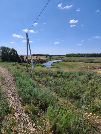 The image depicts a vast, natural landscape under a clear blue sky with scattered white clouds. In the foreground, a gravel path runs along the left side, bordered by lush green vegetation. Two tall, slender concrete poles, part of a power line, stand prominently in the mid-ground, with a single wire stretching diagonally across the sky. Beyond the poles, a winding river meanders through a gently rolling terrain covered in dry grass and interspersed with patches of green. The background features a dense line of trees, forming a treeline that stretches across the horizon. The overall impression is one of a serene, rural environment. The location is identified as Vedenevka, Kazakhstan. There are no people or animals visible in the image.