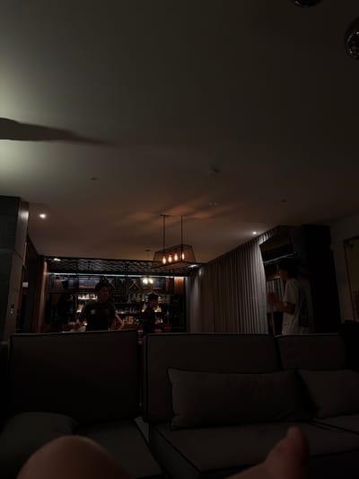 The image shows the interior of a bar or restaurant in Santa Ana Yancuitlalpan (Chilhuacán), Mexico. Several people are present behind the bar, possibly employees or customers. There is a bar setup with bottles of alcohol, indicating it is a serving area. Above the bar, there is a rectangular lighting fixture. The room has a modern design with a dark color scheme and what appear to be metal fixtures. In the foreground, parts of a large sofa are visible, along with what seems to be a person's arm or hand.