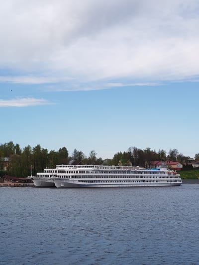 Two white cruise ships are docked side-by-side in the water. Behind the ships is a shoreline with trees and buildings. The sky above is blue with scattered clouds. The water is dark with small ripples. The scene is in Myshkin, Russia.