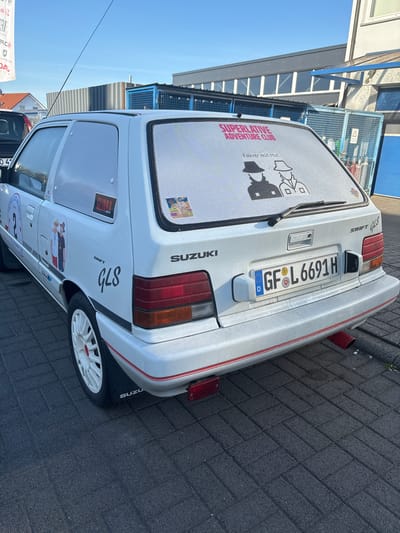 A white Suzuki Swift GLS hatchback is parked on a dark grey brick-paved road. The vehicle has a German license plate "GF•L 6691H." There are several stickers on the car, including one on the rear window that reads "SUPERLATIVE ADVENTURE CLUB" and "Fahrer mit Hut" (Driver with a Hat). The car has white wheels with red accents. The city of Fulda, Germany, is not explicitly identifiable in the image.