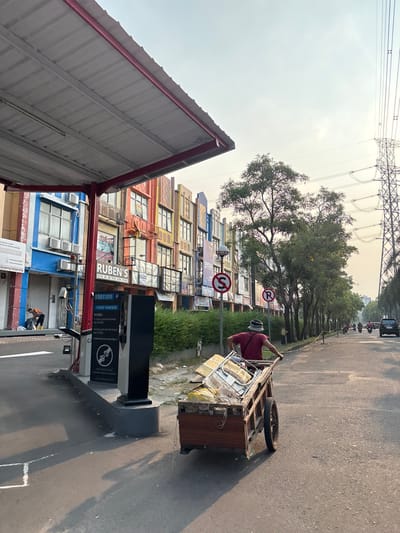 In Pakulonan Barat, Indonesia, a man pushes a wooden cart laden with assorted metal and plastic items down a paved street. He is wearing a maroon t-shirt and a light-colored hat. The cart has two wheels, one of which is larger than the other. The man is walking away from the viewer, towards the right side of the frame.
The scene appears to be in a commercial area with multi-story buildings lining the left side of the street. These buildings have colorful facades and various signage. To the left, a covered structure with a corrugated metal roof extends over a portion of the pavement, under which a parking payment machine is visible. Electrical utility poles and lines are prominent in the background, along with trees and other vegetation. A few motorcycles and a car are also present on the street, suggesting it is an active thoroughfare. The sky is hazy, and the sunlight suggests it is either early morning or late afternoon.