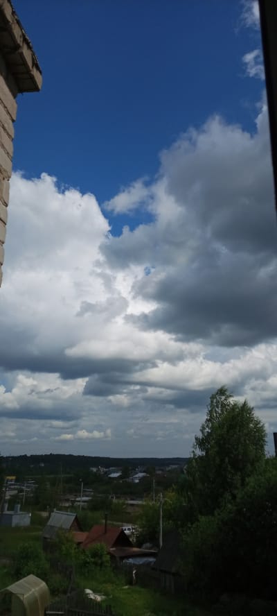 This image captures a view from a building, looking out over a rural landscape under a dramatic sky. The left edge of the frame shows the textured brickwork of the building the photo is taken from, suggesting an elevated viewpoint. The sky is a dominant feature, filled with a mix of bright blue patches and heavy, dark, grey clouds, indicating varied weather conditions, possibly a storm approaching or passing.
Below the sky, a dense line of trees forms a dark green horizon, behind which a scattered village or settlement is visible. Small houses with reddish-brown roofs, some with chimneys, are nestled amongst the trees and open fields. A wooden fence encloses some of the properties. In the foreground, more greenery and what appear to be outbuildings, including a greenhouse structure, are present. A white vehicle is parked near one of the houses. The presence of the greenhouse and the mix of cultivated land and wilder vegetation suggest a rural or semi-rural setting where subsistence farming or gardening might be common.
The lighting suggests it is daytime, likely late afternoon or early evening, given the contrast between the bright sky and the shadowed areas in the landscape. The mood is somewhat foreboding due to the intense clouds, yet also serene with the peaceful village scene below. There is no visible text in the image. The location context provided is Kudymkar, Russia, which aligns with the typical rural architecture and vegetation depicted.