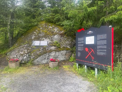 This outdoor media file depicts a memorial site in Elsfjord, Norway, likely related to World War II. The central focus is a large, moss-covered rock formation to the left, with two informational plaques attached to its surface. To the right, a prominent black and red signpost stands, featuring text in both Norwegian and English, along with a map and graphic depictions of a pickaxe and shovel crossed over barbed wire.
The signpost details the history of the "Blodveien" (Blood Road), referencing over 10,000 Yugoslavian prisoners of war brought to Norway between June 1942 and September 1945. They were forced to work on the grand construction of the "Blodveien" and on other infrastructure projects, with many perishing from inhumane conditions, hunger, illness, and mistreatment. The sign also mentions the Lager DASS, where the first prisoners arrived on June 18, 1942, and its subsequent transformation into the Norwegian National Road 50. It notes the closure of Lager DASS on June 18, 1945, and the transfer of prisoners to other locations.
The scene is set in a wooded area, with dense evergreen trees and foliage surrounding the rock and signpost. The ground is covered with gravel and grass, with two potted plants containing vibrant pink flowers placed in front of the rock. The overall atmosphere appears somber and reflective, given the historical context of the memorial. The sky is overcast, suggesting a cool, perhaps damp, day. The time of day is indeterminate, but the lighting is diffused.