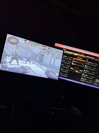 The image shows a laptop with two screens. The lower screen displays gameplay from Counter-Strike: Global Offensive (CS:GO), specifically the "Inferno" map. Player usernames "V3SaKaGOD" and "BOT Rack" are visible, with the former having $5750. The in-game timer shows "0:18". The upper screen displays a social media interface or communication platform with profile pictures and what appears to be match statistics for different profiles.

To the right of the laptop screens, there is a wireless mouse and a mobile phone resting on the laptop's keyboard area. The laptop is identified as an "MSI Stealth 85F RTX" model. The scene is dimly lit, suggesting a low-light environment. The location of the laptop is not determinable from this image. Therefore, no mention of Krumovgrad, Bulgaria, can be included.