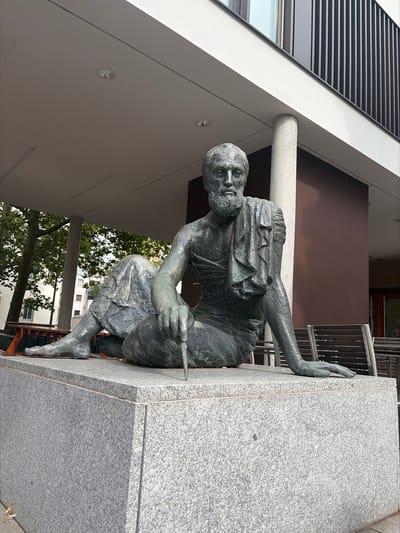 The image shows a bronze statue of a man sitting on a granite pedestal outside a modern building in Würzburg, Germany. The statue depicts a bearded man with his legs crossed, draped in a cloth that resembles a toga. He holds a stylus in his right hand, and his left arm is resting on his bent knee. The man's expression is serious and contemplative.

The statue is located in an outdoor area with a covered walkway supported by a concrete pillar. In the background, there are trees, other buildings, and outdoor seating with chairs and tables. The modern building behind the statue has a dark brown facade and a white upper section with black vertical slats. The sky is visible above the building, appearing overcast. The lighting suggests it is daytime.

The specific location of this statue in Würzburg is in front of the Universitätsbibliothek (University Library) on Am Hubland. This area is part of the university campus.