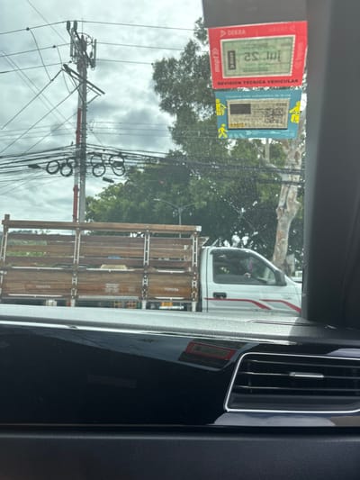The image shows the interior of a car, viewed from the driver's seat, looking out the front windshield. The image is taken in San Rafael, Costa Rica (latitude: 9.937, longitude: -84.152). The windshield is covered with a sticker that reads "REVISION TECNICA VEHICULAR." The sticker is in Spanish and indicates that the vehicle has passed a technical inspection.  A white truck with a wooden bed is visible through the windshield, parked in front of the car. The truck is labeled "TOYOTA."  The truck has a driver in the cab, but their face is obscured by the windshield reflection.  A large telephone pole and numerous power lines are visible behind the truck. There is also foliage in the background. The car dashboard and a vent are visible in the foreground. The sky is overcast.