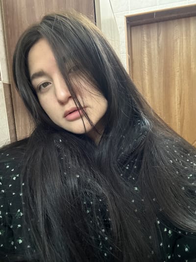 A young woman with long, dark hair lies on a tiled surface, possibly a floor. She is wearing a dark garment with a floral pattern. The background includes a wooden cabinet or drawer. The location is not discernible from the image.