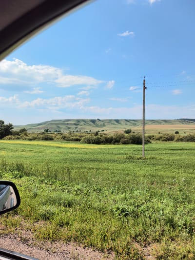 The image depicts a scenic rural landscape under a bright blue sky with scattered white clouds. The foreground is dominated by a lush green field, with some yellow wildflowers visible, hinting at a pastoral setting. A single utility pole with wires stretches across the middle ground, suggesting the presence of infrastructure in this otherwise natural environment.
Beyond the field, rolling hills form the backdrop. These hills are covered in green vegetation, with some areas appearing browner, possibly indicating cultivated fields or drier patches of land. The undulation of the hills creates a sense of depth and vastness.
In the lower left corner, the side mirror of a vehicle is visible, reflecting a portion of the landscape, including the road and the car's body. This perspective suggests the image was taken from a moving vehicle, capturing a fleeting moment of the journey.
The time of day appears to be midday or early afternoon, given the bright sunlight and clear sky. The weather seems pleasant and sunny, conducive to outdoor activities or travel. The overall mood is serene and tranquil, evoking a sense of peace and connection with nature.
The location is identified as Pokrovka, Russia. While there is no visible text in the image, the visual elements strongly suggest a rural area in Russia, characterized by open fields, rolling hills, and utility poles. The scene captures the beauty of the Russian countryside on a clear, sunny day.