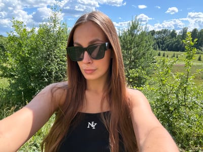 In Шолохово, Russia, a young woman with long, auburn hair, wearing dark sunglasses and a black tank top with a white "NY" logo, is taking a selfie outdoors. She is positioned in the foreground, with lush green foliage and trees forming the background. The sky is a bright blue with fluffy white clouds, suggesting a sunny day. The environment appears to be a natural, possibly rural or park-like setting, with various trees, bushes, and tall grass visible.