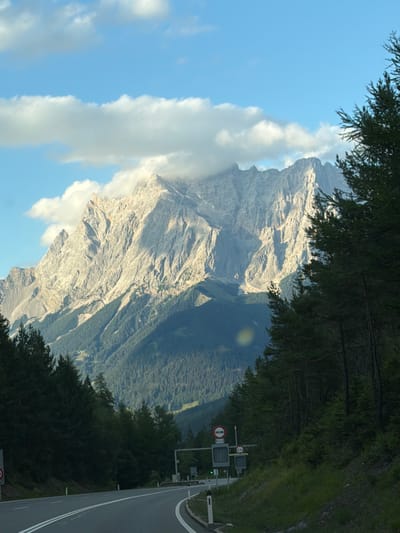 A majestic mountain range dominates the background of this image, its rugged peaks bathed in the warm light of what appears to be late afternoon or early morning. Clouds cling to the higher elevations, adding a dramatic touch to the scene. In the foreground, a paved road curves gently to the right, with white lines marking its lanes. The road is flanked by lush green trees on either side, with the dense evergreen forest on the right side of the frame extending up the mountainside. Along the side of the road, there are traffic signs and a small barrier, suggesting this is a scenic route or possibly a controlled passage. The sky is a vibrant blue, with scattered white clouds that are illuminated by the sun. The overall impression is one of natural beauty and tranquility, with a hint of human presence in the form of the road and its infrastructure.