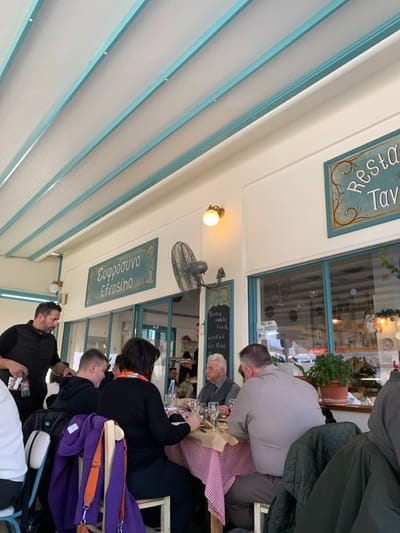 This image, taken in Kastraki, Greece, depicts a lively indoor dining area, likely a restaurant or taverna. The setting features a distinctive ceiling with light blue beams and white panels, possibly part of a covered patio or a skylight structure. Large windows or glass doors separate the dining space, reflecting interior lights and some external elements.
Several people are seated at tables, engrossed in conversation and dining. In the foreground, a bearded man in dark attire is visible, alongside another person with their back to the camera. Further into the scene, a woman with dark hair and an orange lanyard is seated, with an older man with white hair positioned to her right at a table. Another man in a light shirt is seen from behind. Glasses, possibly for wine or water, are on the tables, which are covered with traditional red and white checkered tablecloths. A purple bag hangs casually from a chair.
The walls are decorated with signs, enhancing the local ambiance. One sign at the top reads "Ρεστωράν Ταβ..." (partially visible Greek, likely "Restaurant Tavern"). Another prominent sign below features "Ευφροσυνο Efrosino" in Greek script and Latin transliteration. A smaller board lists menu items: "Home made food," "Cooked," and "Grilled." A "FREE WIFI" sign is visible as a reflection in the window, and a Coca-Cola logo is faintly seen on a potted plant container. A spherical wall lamp emits a warm glow, and a black wall-mounted fan suggests comfort. The overall atmosphere appears casual and welcoming, indicative of a relaxed mealtime.