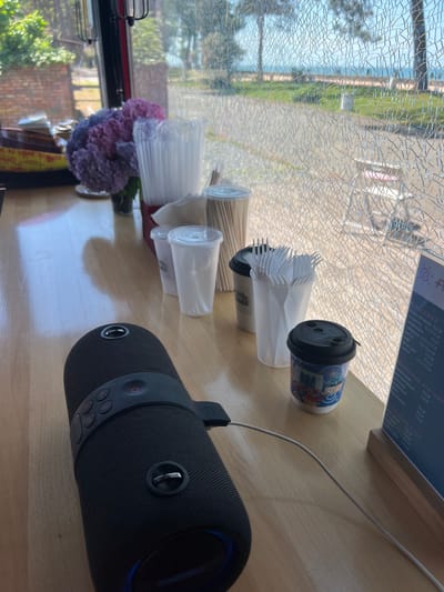 The image shows a close-up view of a wooden counter in what appears to be a cafe or food service establishment. On the counter, a black portable speaker is prominently displayed, connected to a charging cable. To the right of the speaker, several disposable coffee cups, plastic forks, and other disposable drinkware are arranged. A small coffee cup with a colorful design is visible, and a menu board stands to the far right. In the background, through a window with a cracked glass surface, a glimpse of the outdoors is visible, suggesting a location near the sea with trees and a path. The scene evokes a casual, possibly outdoor, dining or takeaway setting, with the overall ambiance suggesting a bright, sunny day. The context of Ochamchire, Abkhazia, places this scene in a coastal town known for its resorts and natural beauty.