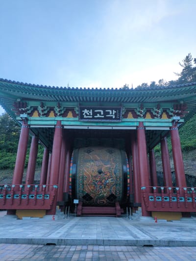 This image depicts the Cheongogak (Heavenly Drum Pavilion), a traditional Korean structure, located in Godang-ri, South Korea. The pavilion is constructed with multiple dark red cylindrical pillars supporting an elaborately decorated, multi-tiered roof featuring green and blue tiles and intricate Dancheong patterns. A black sign with white Korean characters reading "천고각" (Cheongogak) is mounted centrally above the drum within the pavilion.

Inside the pavilion, a very large, cylindrical drum is prominently displayed. The visible face of the drum is adorned with complex, colorful artwork, likely depicting a mythical creature such as a dragon amidst ornate patterns. A decorative wooden railing, painted in red with blue accents, spans the front lower section of the pavilion.

The immediate foreground consists of a light-colored, paved stone plaza, with a visible step leading to a darker, patterned paved area in the far foreground. On the raised platform directly in front of the pavilion, a red fire extinguisher is positioned, along with two silver stanchions. Behind and to the sides of the pavilion, a steep hillside is densely covered with green trees and foliage. The sky above is a clear, light blue. No individuals are present in the frame.