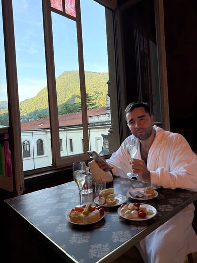 A man wearing a white bathrobe sits at a table in a room with a large window overlooking a mountainous landscape and buildings with red tiled roofs. He is holding a glass of champagne and a smartphone. On the table are several small plates of food, including cheese, meat, and pastries, as well as glasses of champagne and water. The scene appears to be set in San Pellegrino Terme, Italy, given the architectural style and the presence of what looks like a historic building and the mountainous backdrop characteristic of the region. The man is looking towards the camera with a relaxed expression.