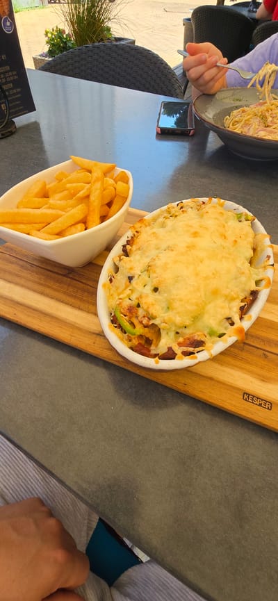 The image shows a table setting at an outdoor restaurant in Haren (Ems), Germany, as indicated by text on a menu visible in the upper left corner. On the table, there are two dishes of food: a white oval dish filled with French fries and a white oval gratin dish topped with melted cheese, containing what appears to be a casserole or baked pasta dish with visible green peppers and other ingredients. A wooden cutting board is placed beneath these dishes. In the background, a person's hand is visible holding a fork and interacting with a bowl of spaghetti carbonara. A smartphone is also present on the table, along with dark woven chairs and some greenery. The overall scene depicts a meal being served and consumed outdoors.