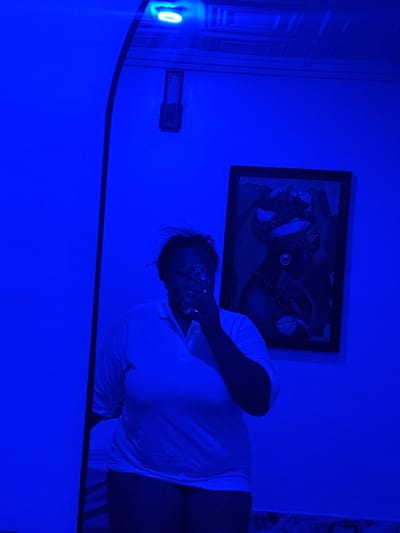 The image shows a person lying on a bed in a room with a blue light. A framed picture hangs on the wall behind the person. The person is holding a phone in front of their face. The location is a room in Rumuigbo, Nigeria.