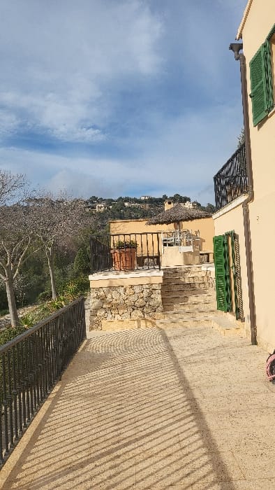 This image depicts an outdoor, sunny, terraced residential area within the Polígon Industrial De Calvià, Spain. The foreground features a light-colored, possibly stone-tiled, paved pathway extending from the lower left to the upper right. A dark metal railing runs along the left edge of this pathway, casting prominent parallel shadows onto the pavement, indicating strong direct sunlight.
To the right, a light-beige building with a visible white downspout and two closed green louvered shutters is present. Adjacent to this building, the pathway transitions into a series of wide, light-colored stone steps ascending towards the midground.
In the midground, a stone retaining wall supports an elevated terrace. On this terrace, a dark metal railing borders an area where a large brown terracotta pot with visible foliage is situated. Behind this, a second light-beige building features another elevated terrace topped with a distinctive thatched umbrella or parasol, beneath which white outdoor furniture, including chairs and a table, is visible.
The background consists of a sloping landscape with numerous trees, some deciduous with bare branches, and verdant green foliage. Hills dotted with residential structures are visible in the far distance under a bright blue sky with scattered white clouds. No individuals are present in the scene.