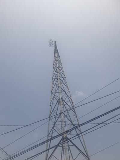 The image shows a tall, lattice-style telecommunications tower against a hazy, light blue sky. Several power lines crisscross the foreground and middle ground, some passing in front of and behind the tower. The tower itself is constructed of metal beams forming a triangular structure that tapers towards the top. At the very apex of the tower, there are three circular antenna elements mounted horizontally. The sky appears overcast or possibly dusty, with a faint, diffused light. No people are visible in the image. The scene suggests an outdoor, urban or semi-urban environment, common in areas like Watesalit, Indonesia, where infrastructure for communication and power distribution is prevalent.