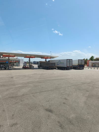 This image captures a vibrant daytime scene at a Galp service station in Torrent, Spain. The setting features a large paved area with several vehicles and a prominent gas station structure under a bright blue sky dotted with scattered white clouds.
In the foreground, the expansive asphalt surface shows some tire marks and patches of wear. To the left, the main gas station canopy, branded "Galp" in red and white, covers multiple fuel pumps. A person in a light shirt is seen near a motorcycle or scooter parked at one of these pumps. Another person is partially visible behind a red car, which is also positioned at a pump. The gas station building, with glass doors and a visible disability access symbol, stands behind the pumps.
To the right of the gas station canopy, a large grey truck with an open-top trailer (possibly a dump truck or bulk carrier) is parked, extending towards the right side of the frame. Further right, two white semi-trailers are parked side-by-side. The left-most white trailer clearly displays the text "TTES EDY SPEDITION S.L." and "(GANDIA, VALENCIA)," indicating its origin or area of operation. The second white trailer also has a logo and text, though less discernible. In the background on the far right, a car wash facility, indicated by the word "lavado," is visible, partially obscured by trees.
The activity suggests a typical service stop, with vehicles refueling or taking a break. The bright sunlight indicates it is likely mid-day or early afternoon. The presence of several large trucks points to this being a significant stop for commercial vehicles.