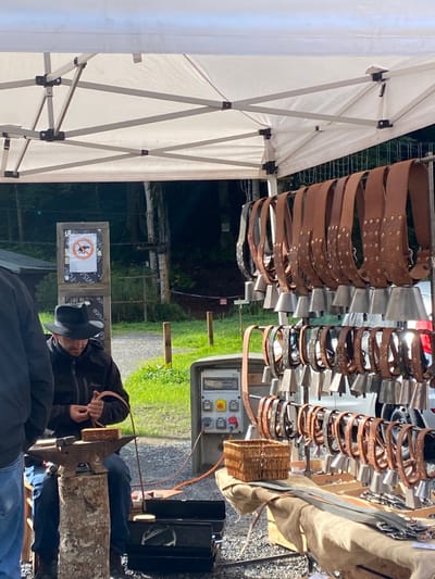 The image captures an outdoor market stall or craft demonstration situated in Abondance, France. Under a white fabric canopy supported by a metal framework, a male individual wearing a dark fedora-style hat, dark zippered jacket, and blue jeans is seated on a stool at the left. He is actively engaged in crafting, manipulating a piece of brown leather with both hands over a small, dark-colored anvil that rests on top of a vertical wooden log acting as a stand. Another individual's left torso and arm are partially visible on the extreme left, facing towards the craftsman.
To the right of the craftsman, a multi-tiered display rack holds numerous brown leather belts, each adorned with multiple metal bells of varying sizes, commonly known as cowbells. These belts are arranged horizontally across several metal rods. Below this primary display, additional brown leather straps and metal buckles are laid out on a light-colored fabric covering a lower surface. A small, light-brown wicker basket is positioned on this surface. Behind the display, an industrial-style grey electrical control panel with a top-mounted gauge, red circular buttons, and yellow toggle switches is visible, with an orange power cord extending from it towards the left.
The background reveals an outdoor setting: a light-colored gravel path extends from left to right, bordered by green grass and sparse wooden posts. Beyond the path, a dense, dark-green wooded area with tall trees forms the backdrop. A square wooden sign depicting a red circle with a diagonal line over a dog icon, signifying "no dogs allowed," is mounted on a wooden post near the path. The scene is illuminated by natural light, indicating daytime.