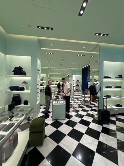 The image displays the interior of a luxury retail store, identified as a Prada boutique located within Paris Charles de Gaulle Airport (CDG) in Roissy-en-France, France. The environment features mint green walls and ceiling, illuminated by recessed lighting and shelf strip lights. The floor is composed of large, polished black and white marble tiles arranged in a checkered pattern.
In the mid-ground, two men are engaged in an interaction near a glass-topped display counter. The man on the left, with a short beard, wears a dark long-sleeved shirt and light grey trousers, facing right towards the second man. The man on the right, with short dark hair, wears a light-colored short-sleeved shirt and dark trousers, standing behind the counter and appearing to assist or serve the first individual. The counter displays small items, possibly watches or accessories. A third, partially visible person is further in the background to the left.
The store is furnished with floor-to-ceiling shelving units along the walls. On the left, these shelves display various black leather and nylon bags, including backpacks, a duffel bag, and a cap, along with other small items. On the right, shelves showcase multiple pairs of men's shoes, ranging from sneakers to dress shoes and boots. A white torso mannequin, positioned on the right side, displays a black backpack. Adjacent to this mannequin, a black rolling suitcase with an extended handle stands upright on the floor. In the left foreground, a dark green upholstered ottoman is visible next to a lower glass display case containing wallets and cardholders.