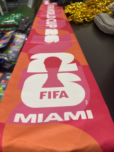 A high-angle, close-up photograph, taken in Sunrise, United States of America, captures a rectangular fabric item, likely a scarf or banner, displaying FIFA World Cup 2026 branding. The item lies diagonally across a dark, smooth surface from the bottom-left to the top-right of the frame. Its vibrant design features geometric shapes in various shades of pink and orange. Prominently displayed in the foreground is the word "MIAMI" in white capital letters on a dark pink panel. Above this, the word "FIFA" is rendered in dark pink capital letters, set against a white silhouette of the number "26," which also incorporates a dark pink graphic representation of the FIFA World Cup trophy. Further along the fabric, partially visible and inverted, is the text "FIFA WORLD CUP 26" in white. To the left of the fabric item, out-of-focus background elements include stacks of green and white patterned merchandise, a multicolored item with a square pattern, and plastic packaging, resting on what appears to be a green carpet or fabric. To the top-right, also out of focus, are a pile of shiny gold, tinsel-like material and a folded light gray fabric item, all on the dark surface. The central fabric item is in sharp focus, while the peripheral objects are blurred.