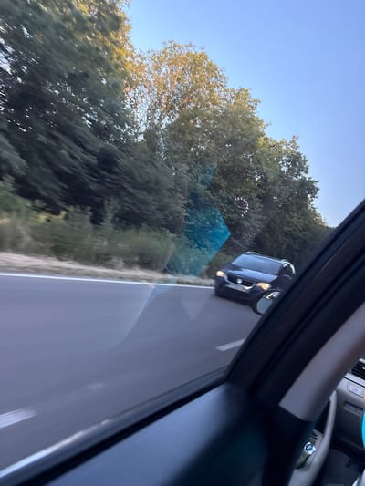 The image, taken from inside a car, shows a dark-colored SUV driving on a road. The SUV is in the left lane, and the view is from the passenger side, looking forward and slightly to the right. Trees line the right side of the road, and the sky is clear blue, suggesting a sunny day. The angle of the shot is skewed, as if the photo was taken with the camera held at an unusual angle or while the car was moving. The interior of the car is partially visible in the foreground, with a black pillar and part of the dashboard.
A faint, circular image of a speedometer can be seen reflected on the windshield, indicating the car's speed. The trees are lush and green, indicating it is likely summer or late spring. The lighting suggests it is either early morning or late afternoon, with the sun casting a warm glow. The image appears to be taken in a rural or suburban area, given the presence of trees and the open road. There is no visible text in the image. The overall impression is a snapshot of a journey, capturing a moment of travel through a scenic route.