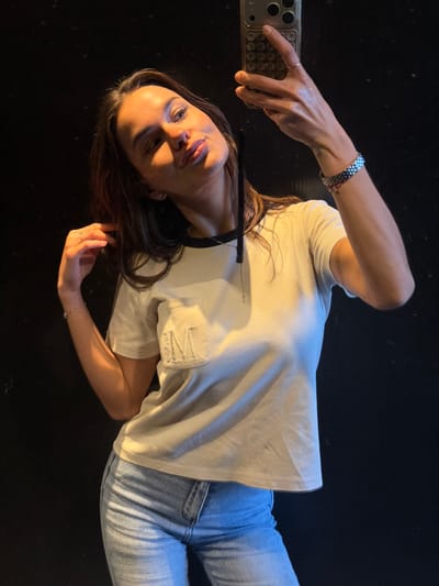 A female subject is depicted from the upper thighs upwards, captured in a mirror selfie in an unspecified indoor location within Pardes Hana - Karkur, Israel. She holds a smartphone, possibly an iPhone, in her right hand to take the photograph. Her left hand is positioned near her left shoulder, touching her hair or neck. The subject is wearing a light-colored, short-sleeved t-shirt with a black collar trim and a left chest pocket embroidered with the letter "M." She is also dressed in light blue denim jeans. On her right wrist, a multi-colored (blue, silver, black) bracelet is visible, and a thin, light-colored bracelet adorns her left wrist. A ring is present on her left ring finger, and a thin necklace with a pendant, along with a black strap, is around her neck. The background is uniformly dark, providing no discernible environmental details other than the reflective surface in which the selfie is captured. The subject's head is tilted slightly, and her lips are pursed towards the camera.