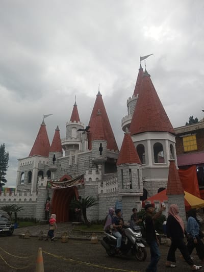 The image displays a large, elaborate castle-themed building under a heavily overcast, grey sky in Suddepalle, India. The structure is constructed with grey, stone-textured facades, featuring multiple red-tiled conical and pyramidal roofs, fortified battlements, and arched openings. Several small white flags are visible atop various spires. A dark, winged dragon-like figure is mounted on a central tower. A colorful banner is suspended above the main arched entrance.
In the foreground, a paved ground surface exhibits orange traffic cones and yellow tape or rope. Multiple individuals populate the scene. A silver motor scooter, possibly a Yamaha NMAX, is occupied by three people: a driver in a light blue shirt, a child positioned in front of the driver, and an adult wearing a purple hijab seated behind, holding a small child. Adjacent to the scooter, a young male in a black t-shirt and blue jeans walks forward, holding a smartphone aloft. To the right, a person in a dark abaya and pink hijab walks away from the castle, followed by another individual in a light blue jacket and dark pants. Near the castle's main entrance, a small child in pink walks on the paved path.
To the far left, a black SUV is partially visible, alongside a tall coniferous tree. Small palm trees are planted around the base of the castle, near decorative bollard-like structures. To the right of the castle, a section of another building with a brown brick facade and a distinct yellow-framed window is visible, partially obscured by orange and yellow fabric awnings or tarps.