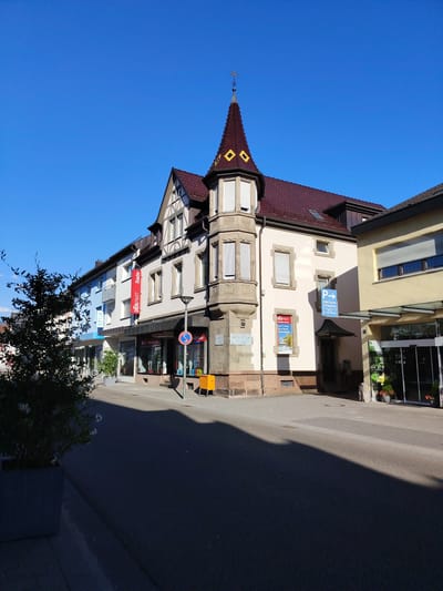 This is a landscape, eye-level, outdoor photograph taken on a clear, sunny day in Achern, Germany. The image features a row of commercial buildings lining a street. The building in the foreground on the right is a modern structure with large glass doors and windows, likely a shop or cafe. To its left, an older building with a distinctive turret topped by a decorative spire stands prominently. This building showcases a mix of architectural styles, with a half-timbered upper section and a stone base. A red banner with German text, "DERPART Rade," hangs from the facade of one of the buildings. Other visible text includes "OPTK," "P STADTCAFE," and "40 Parkplätze," indicating businesses and parking information. The street in front of the buildings is paved, and a large, dark shadow from a tree and potentially another object falls across the lower portion of the image. A potted tree with sparse foliage is visible on the left side of the frame. The sky is a vibrant, cloudless blue, suggesting it is daytime. The overall scene depicts a typical European town street with a mix of old and new architecture, and commercial activity. No people are clearly visible in the frame.