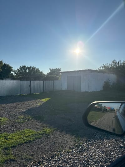 This is a sideways, eye-level outdoor, handheld shot captured on a bright, sunny day. The sun is positioned in the upper left quadrant of the frame, casting a strong lens flare and bright beams across a clear blue sky. In the foreground, the scene is dominated by a gravel driveway. To the right of the driveway, lush green grass grows sparsely between the stones, and a dark shadow is cast by an unseen object.
Dominating the middle ground is a long, low-slung building with a white, slightly weathered facade. In front of the building, and running parallel to it, is a row of what appear to be corrugated metal panels or structures, stacked in a segmented fashion. To the left of this structure, more trees are visible, their green foliage contrasting with the bright sky.
On the far right of the frame, a car's side-view mirror is visible, reflecting a scene of the road ahead with various vehicles and structures. The reflection shows a road, more gravel, and what appear to be colorful buildings in the distance. The overall impression is of an industrial or utilitarian area, possibly on the outskirts of a settlement, given the presence of the gravel, buildings, and what looks like fencing. The time of day is clearly daytime, with the sun high in the sky, indicating it is likely mid-morning to early afternoon. The weather appears to be clear and fair. There is no visible text in the image.
