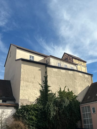 This image captures a multi-tiered architectural scene in Weißenburg in Bayern, Germany, under a bright blue sky with scattered cirrus clouds. The dominant feature is a massive, light-beige or cream-colored textured wall, possibly a foundation or retaining wall, rising steeply from the foreground. This wall exhibits a dark horizontal trim line near its top and several subtle rectangular indentations. Extensive dark green ivy and other dense foliage cover the lower and mid-sections of this wall, with prominent tendrils ascending vertically.
Above and behind this primary wall, a complex of buildings is visible. To the left, a light-beige plastered building with a dark brown gabled roof and a small rectangular window is present. Further to the right, a flatter, lower section with multiple windows and a slight overhang is seen. The uppermost and furthest right structure is a distinct half-timbered (Fachwerk) building with dark timber beams against a lighter, possibly yellow, infill, topped by a dark gabled roof.
Partial views of other structures include a dark tiled roof on the far lower-left and a pink-hued building with a large grid-patterned window and a smaller white-framed window on the far lower-right. The scene is well-lit by direct sunlight, creating clear shadows. No individuals or moving objects are present.