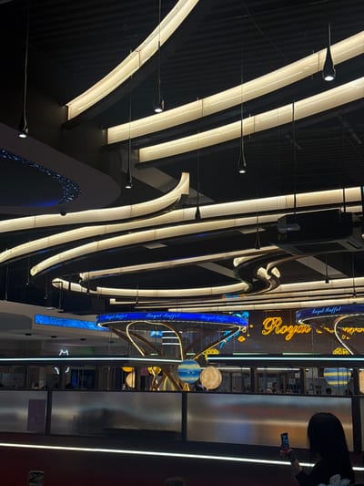 The image shows a modern interior with a dynamic display of linear and curved lights, creating a striking visual pattern against a dark background. A "Royal Buffet" sign is prominently featured, adorned with decorative elements. Reflective surfaces and a glimpse of the restaurant’s interior are visible on the right. People are partially seen, adding a sense of scale and activity to the scene.