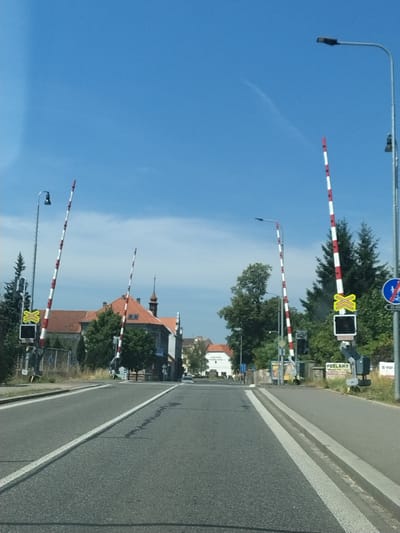 This media file depicts a road scene in Horažďovice, Czechia, under a clear blue sky, suggesting fair weather and daytime. The primary subjects are several railway crossing barriers, each marked with alternating red and white stripes. These barriers are raised, indicating that a train is not currently approaching or has already passed. One barrier, in the foreground, has a yellow 'X' sign and a black square light. Further down the road, another similar barrier is visible.
In the background, there are buildings with red tile roofs, characteristic of European architecture, and lush green trees. A road with visible white lane markings runs alongside the railway crossing. The scene appears to be a typical intersection of a road and a railway line. No people are visible in the frame.
Some text is discernible on signs: a circular blue sign with a white symbol (likely a traffic or railway warning) is present at the top of the frame. Another rectangular sign with white text on a dark background, reading "PODLAHY" and "PE." is visible next to a barrier, and a smaller blue traffic sign is also present. The overall impression is one of a quiet, ordinary day in a town.
