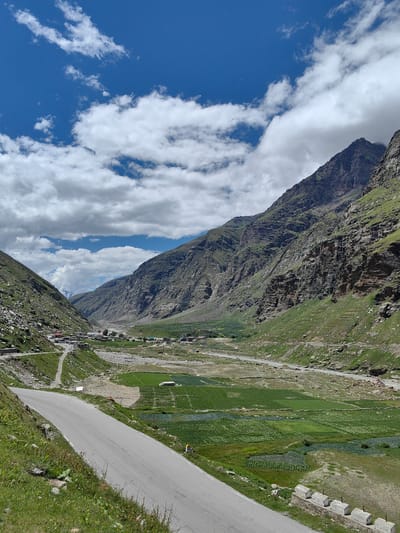 The image shows a wide, paved road curving through a mountain valley in Koksar, India. Steep, rocky, and grassy mountainsides rise on either side of the valley. The valley floor is a patchwork of cultivated fields, some with crops appearing to be in rows, and a meandering river. In the distance, a small village or settlement is visible nestled in the valley. The sky is blue with fluffy white clouds. The road is empty of vehicles. The overall scene depicts a remote, natural landscape with evidence of human habitation and agriculture.