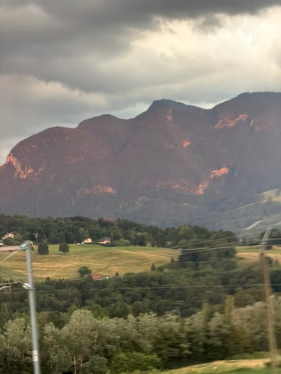 The image shows a mountain range in the background with a cloudy sky above. The mountains are covered in trees and some areas are illuminated with a reddish-orange light, suggesting the sun is setting or rising. In the middle ground, there are rolling hills with green fields and scattered houses and trees. In the foreground, there is a line of trees and a power pole with wires running across the frame. The scene appears to be a rural landscape, possibly in the vicinity of Groisy, France.