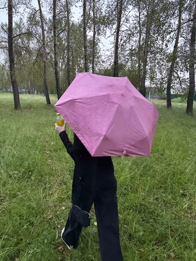 In a grassy area with trees in the background, a person is holding a pink umbrella over their head. They are wearing black pants and shoes. The person is also holding a clear plastic bottle with a yellow liquid in it. The sky is overcast, suggesting it may be raining or cloudy. The trees have light-colored bark and are mostly bare of leaves, indicating it might be autumn or early spring. The grass is tall and green, with some patches of dried leaves visible. The overall scene suggests a casual outdoor activity. There is no visible text in the image. The location context provided is Nevyansk, Russia.