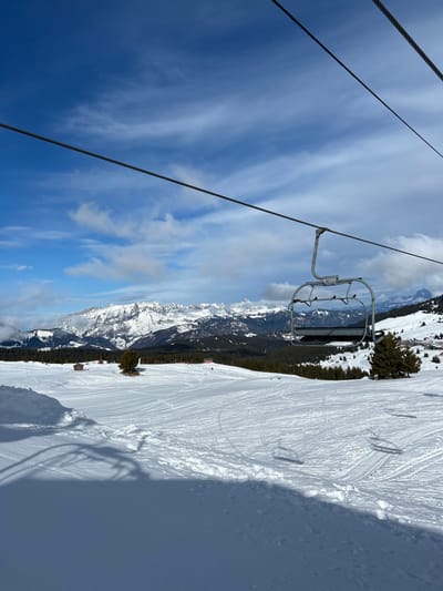 The image displays a bright, snow-covered mountain landscape, identified as a ski area in Villard-sur-Doron, France. The foreground consists of white snow, marked by various ski tracks and a prominent dark shadow extending from the bottom left. Two dark, parallel overhead cables stretch horizontally across the upper half of the frame. Suspended from the lower cable is an empty, grey metal ski lift chair, visibly numbered "11" on its safety bar.
The midground features a wide, groomed ski slope blanketed in snow, showing numerous parallel ski tracks. Several small, dark wooden structures, resembling huts or cabins, are visible on the left side of the slope, along with a few scattered evergreen trees. Red boundary markers are sparsely distributed across the slope.
In the background, a range of large, rugged snow-capped mountains dominates the horizon, partially obscured by low-lying white clouds clinging to their peaks. Below the mountain range, dark green coniferous forests are visible. The sky is a vibrant blue with scattered white, wispy cirrus clouds. No individuals are visible on the ski slope or within the ski lift chair.