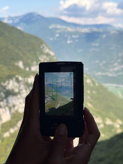 A hand, adorned with a thin gold ring on the ring finger, holds a black digital camera in front of a blurred, expansive mountain landscape. The camera's screen displays a grid overlaying a view of the same mountain vista, indicating it is actively framing a shot. The background features rolling green hills and distant, hazy mountains under a bright sky with scattered clouds. The scene suggests a moment of capturing the natural beauty of the mountainous terrain, likely in a location like Ferrara di Monte Baldo, Italy, known for its scenic views.