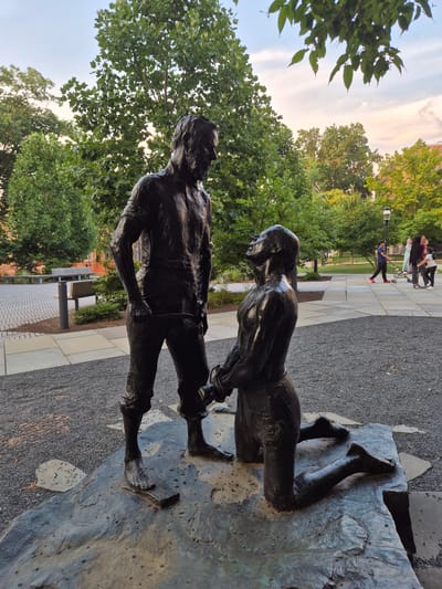 The image depicts a bronze sculpture of two men in an outdoor setting. The sculpture is positioned on a rough, dark stone base. One man stands with his hands in his pockets, looking down at the other man who is kneeling. The kneeling man has his head tilted back and his hands appear to be bound together in front of him. Both figures are muscular and appear to be wearing rough clothing, possibly representing laborers or historical figures.

The sculpture is located in Princeton, New Jersey, USA, in a plaza area. Behind the sculpture, there is a paved walkway with a patterned brick design. Several benches are visible in the background, along with lush green trees. In the far background, to the right of the sculpture, several people are walking and one individual is on a scooter. The sky is visible above, with a soft blend of orange, pink, and blue hues, indicating it might be either dawn or dusk. The ground surrounding the sculpture is covered in dark gravel.