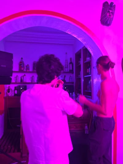 An indoor scene in Fortín de las Flores, Mexico, is illuminated by pervasive magenta and purple lighting, creating a vibrant atmosphere. Two individuals are present within a room featuring a prominent arched doorway. In the foreground, a male, seen from behind, has dark, curly hair and wears a light-colored short-sleeved shirt. His right hand is raised near his neck. To the right of the frame, a female is depicted in profile, facing left, with her hair pulled into a bun. She appears shirtless and wears dark cargo-style pants, with her arms bent and hands in motion, suggesting a dynamic pose or dancing.
The environment includes a bar or counter area behind the male, equipped with various liquor bottles, including a discernible Johnnie Walker Red Label bottle, and multiple mugs or glasses. Two dark-colored speakers are positioned on the counter. Beyond the female, to the right of the archway, a wooden shelving unit displays bottles, likely wine. The archway itself is adorned with a red or orange trim, and a carved wooden mask with a stylized face is mounted on the wall above it, to the right. The overall setting appears to be an entertainment space or bar.