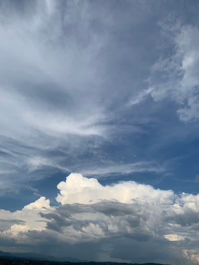 The image captures a dramatic sky over the city of Khust, Ukraine. The foreground is dominated by a vast expanse of clouds, ranging from bright white, puffy cumulus clouds to darker, more ominous storm clouds. These clouds fill the majority of the frame, creating a sense of depth and scale. The upper portion of the image transitions into a deep blue sky, with wispy cirrus clouds streaking across it, illuminated by sunlight. In the far distance, a hazy silhouette of mountains and hills is visible, with faint outlines of buildings and structures suggesting the presence of the city of Khust. The overall impression is one of a dynamic and changing weather system.