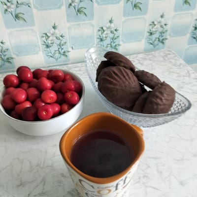 A medium shot captures a still life arrangement on a marble-patterned countertop. To the left, a white bowl overflows with ripe, red cherries. In the center, a clear glass bowl holds several chocolate-covered cookies, their rippled texture visible. In the foreground, an orange-rimmed mug filled with dark liquid, likely coffee or tea, is positioned to the right. The background features a light blue wallpaper with a repeating floral pattern of white flowers and green leaves, suggesting an indoor setting. The scene appears to be set during daylight, given the ambient lighting. The arrangement evokes a sense of a quiet moment, perhaps a break or a simple breakfast. Visible text on the mug reads "Coffee Time".
