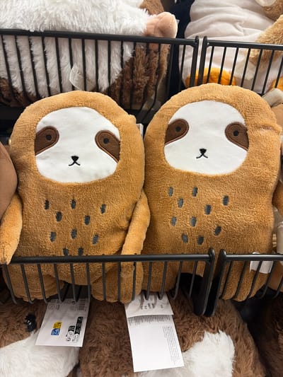 Two plush sloth toys with brown fur and white faces are displayed in a black metal rack. The toys have dark brown eye patches and small black embroidered mouths. Their bodies are covered in dark grey spots. The background shows other plush toys and shelves in a retail environment. One tag visible on the rack has recycling symbols and text in multiple languages. The scene appears to be a retail store, possibly a toy store or department store, located in Würzburg, Germany.