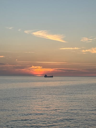 A cargo ship is visible in the distance on a calm sea at sunset. The sky is painted with a gradient of soft blues, oranges, and pinks from the setting sun, with wispy clouds scattered across it. The water reflects the colors of the sky, creating a serene and peaceful atmosphere. The ship appears to be heading towards the horizon, adding a sense of journey and tranquility to the scene.