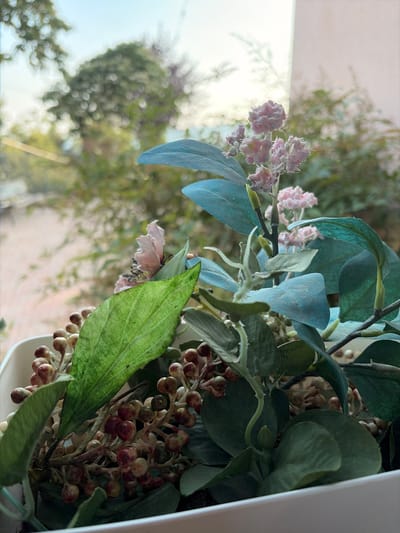 This is a close-up, eye-level, vertical shot of an artificial floral arrangement placed near a window. The arrangement features a variety of elements, including large, muted blue-green leaves, smaller pinkish-purple clusters of flowers, and branches dotted with small, bead-like berries in shades of cream, burgundy, and green. A prominent, textured green leaf with visible veins is positioned in the foreground, slightly to the right of center.

The background is softly blurred, suggesting an outdoor scene viewed through the window. Hints of trees and foliage, bathed in bright, diffused light, indicate it is daytime, likely sunny, with clear skies visible. The soft focus and the interplay of light and shadow on the artificial flowers and leaves create a gentle, somewhat serene atmosphere. The arrangement appears to be indoors, possibly in a home or a commercial space in Cornedo Vicentino, Italy, given the provided location context. There is no visible text in the image.
