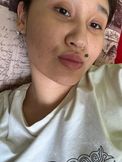 The image is a close-up, overhead shot of a young woman lying down, likely in a bedroom in Koçkor-Ata, Kyrgyzstan. She is wearing a light-colored t-shirt and has a mole on her right cheek. Her hair is dark and pulled back. She is wearing a small diamond earring in her left ear. The background includes a patterned pillow with script and a red textured surface, possibly a blanket or another pillow. The lighting suggests an indoor setting, possibly natural light from a window.