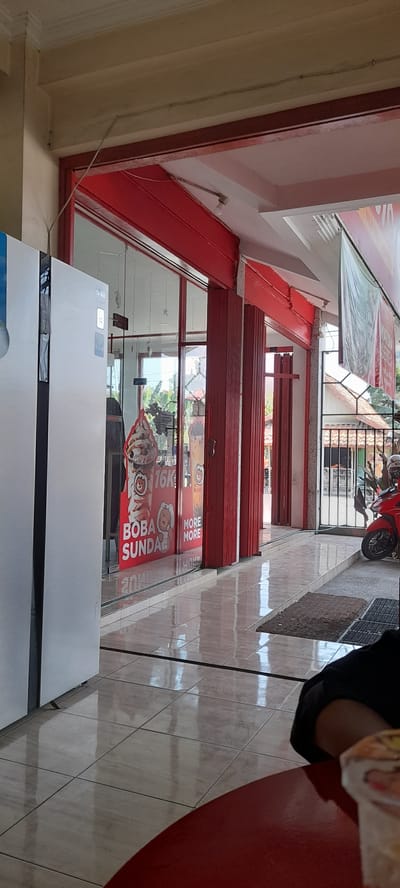 The image depicts the exterior of a shop with red folding doors and large glass windows. A white refrigerator is visible on the left, and advertisements for "BOBA SUNDAE" and "MORE MORE" are displayed on the glass. Inside, there are some hanging decorations. The floor is tiled and reflective. A red motorcycle is parked outside on the right, next to a gated area. The scene appears to be in an outdoor commercial area, possibly a street in Crewek, Indonesia. A person's arm and shoulder are visible in the foreground on the right, resting on a red circular surface, possibly a table.