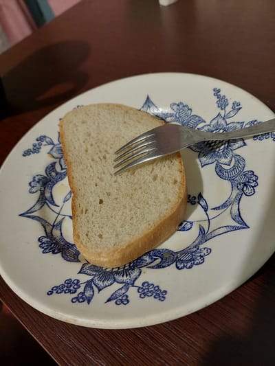 A close-up shot shows a slice of white bread on a white plate with a blue floral pattern. A silver fork is embedded in the bread, with its tines visible. The bread appears to be a simple loaf, with a light brown crust and a soft, airy interior. The plate has a decorative border with intricate blue floral designs, typical of traditional Ukrainian ceramics. The background is dark and out of focus, suggesting the photo was taken indoors, possibly on a wooden table. The lighting appears to be artificial, casting a shadow on the left side of the plate. The image is clear and well-lit, highlighting the texture of the bread and the details of the plate. There is no visible text in the image. The setting suggests a casual meal or snack.